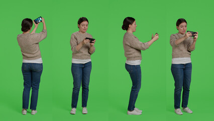 Female model playing video games online on smartphone, standing over greenscreen backdrop and having fun. Young adult enjoying shooter gaming competition play in studio, positive woman.