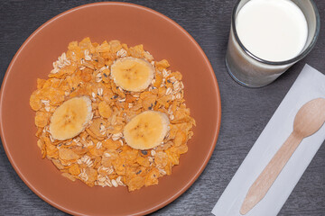 A bowl of corn cereal with raw oats, chia seeds and three slices of banana next to a glass of milk and a spoon on a wooden table