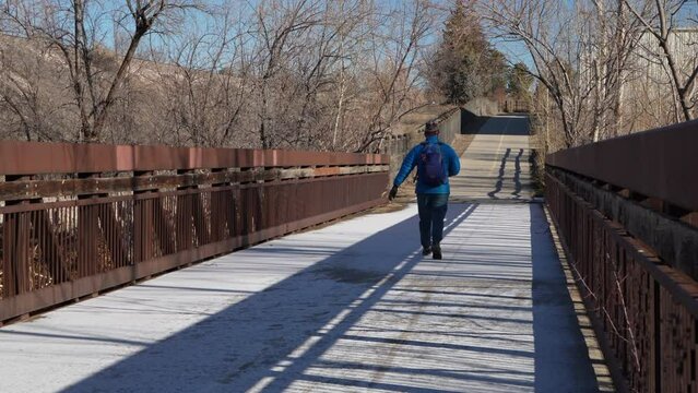 older man with a small backpack is walking on a bike trail and footbridge covered by frost in Fort Collins, Colorado, winter morning
