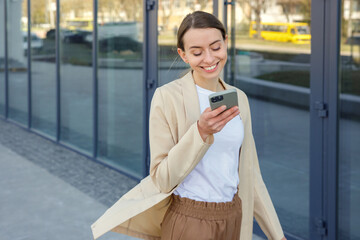 Happy caucasian woman with smartphone in hand going to record voice message to colleague outdoors. Smiling female employee in official wear walking near business center.
