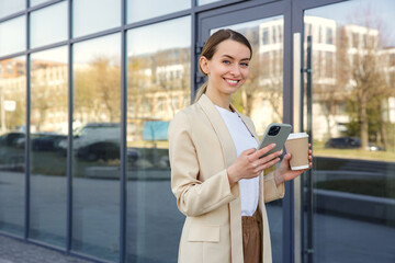 Portrair of the busy caucasian business woman walking, using smartphone and holding coffee cup on street. Attractive female typing message near office building during coffee break.