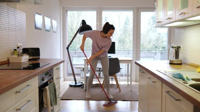 Active girl with headphones, dancing at home while cleaning with a mop in her hands, relieves stress and lifts the mood.