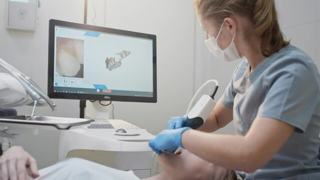Doctor scans the patient's teeth in the clinic. The dentist holds in his hand a manual 3D scanner for the jaw and mouth. Dental health. Creates a 3D model of teeth and gums on a medical monitor.