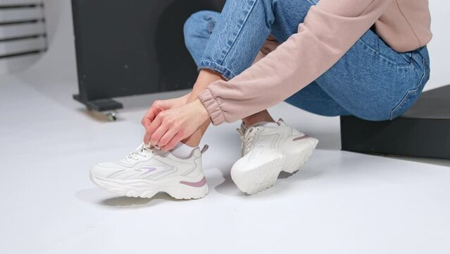 Lady in jeans and sweatshirt ties the laces on her white sneakers. Girl in modern look poses in studio.