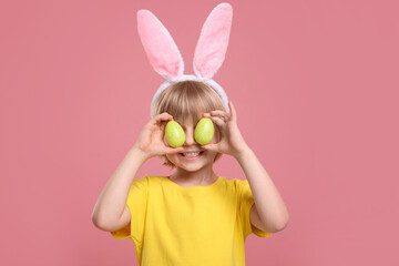 Happy boy in bunny ears headband holding painted Easter eggs near his eyes on pink background