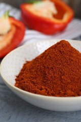 Bowl with aromatic paprika powder and fresh bell peppers on grey wooden table, closeup