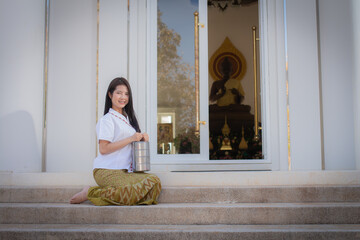 Beautiful thai woman in a white traditional thai dress is sitting while holding a lunch box called a pinto in front of a white temple.
