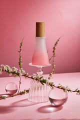 Scene mockup for cosmetic product with empty bottle without label on glass cup, transparent balls and flower branches on pink background. Front view, minimal concept, beauty product design.