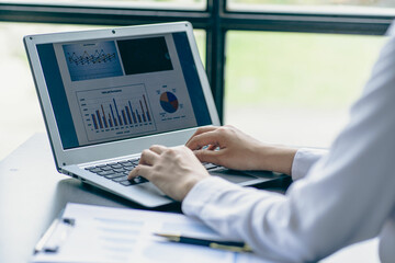 Smiling Asian businesswoman working on laptop computer at her desk in modern home office doing calculations analyzing expenses reporting finances