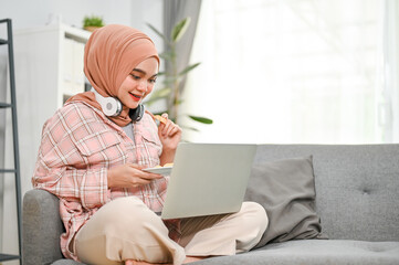 Happy young Asian Muslim woman eating cookies and watching movie on her laptop