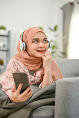 Cheerful Asian Muslim woman relaxing in her living room, listening to music on sofa.
