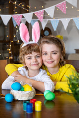 Obraz premium Easter Family traditions. Two caucasian happy children with bunny ears dye and decorate eggs with paints for holidays while sitting together at home table. Kids embrace and smile in cozy. Vertical