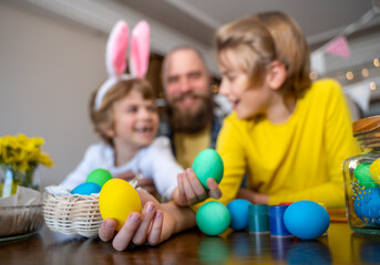 Fototapeta premium Easter Family traditions. Father and two caucasian happy children with bunny ears dye and decorate eggs with paints for holidays while sitting together at home table. Kids embrace and smile in cozy