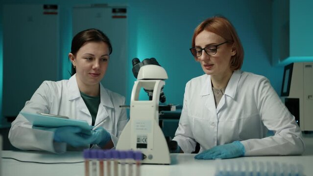 Women in lab coats looking through a microscope and writing something. They are in a laboratory performing an important sample analysis. They are highly skilled professionals High quality 4k footage 