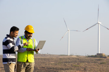 India engineer using laptop and showing control of windmill farm operation to generate electricity, Asian man wearing yellow helmet and vest working at wind turbine farm, clean and green energy.