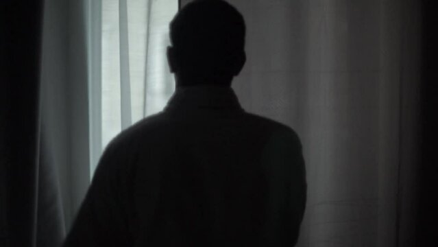 Silhouette of a mysterious and stalker-like man opening the curtains of the window of his house to look outside