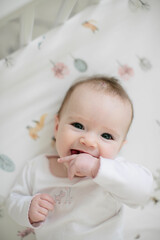 sweet caucasian baby girl smiling with hand in her mouth looking up at the camera from patterned crib sheet