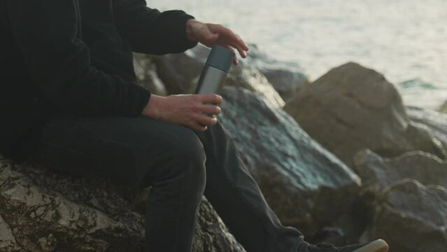 man unscrews thermos and pours hot drink into cup as sea waves wash over rocks in background during golden hour