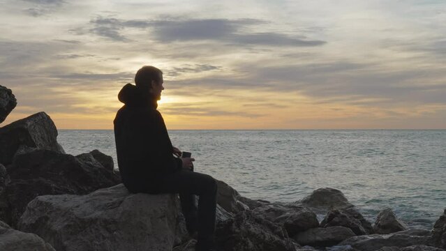 man enters, sits on a rock, opens thermos, pours hot  drink and enjoys sea view during golden hour