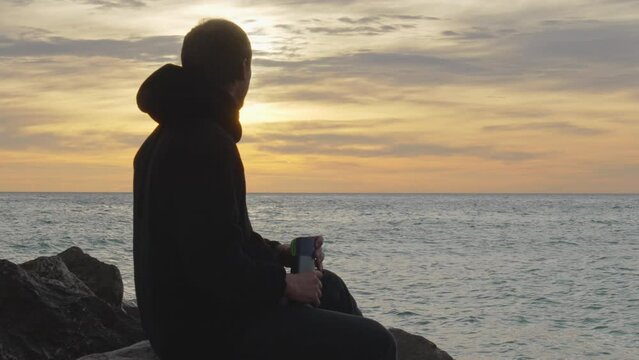 man enters, sits on a rock, opens thermos, pours in, drinks and enjoys the sea view during golden hour