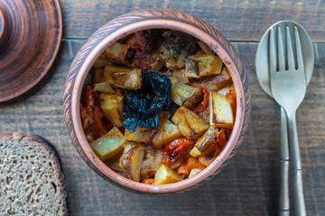 Stewed potato, carrot, onion, tomato and prunes in a clay pot on wooden background, closeup