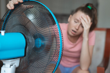 Young woman using electric fan at home in living room, sitting on couch cooling off during hot...