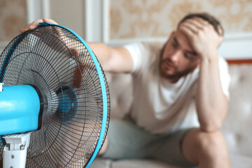 Young bearded man using electric fan at home, sitting on couch cooling off during hot weather, suffering from heat, high temperature - obrazy, fototapety, plakaty