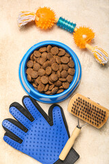 Bowl of dry pet food, toy and grooming brush on light background, closeup