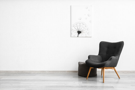 Stylish grey armchair, pouf and painting with dandelion near white wall