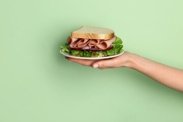 Female hand holding plate with delicious ham sandwich on green background
