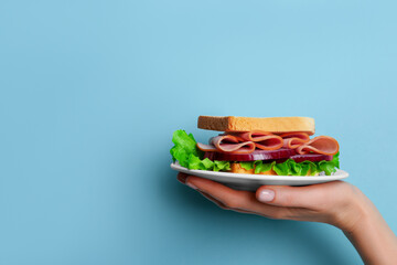 Female hand holding plate with delicious ham sandwich on blue background