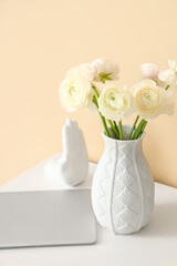 Vase with ranunculus flowers and laptop on table near beige wall, closeup