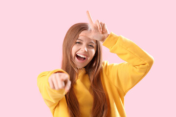 Young woman showing loser gesture and pointing at viewer on pink background, closeup