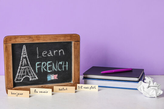 Chalkboard with text LEARN FRENCH, words and books on table near lilac wall