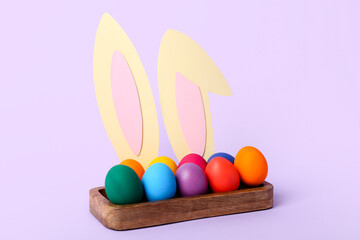Box with Easter eggs and bunny ears on lilac background