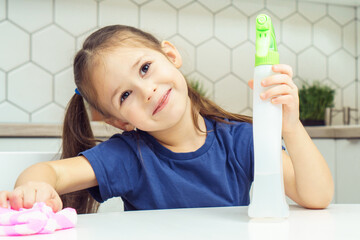 Happy little girl with detergent sprayer and household rag at table. Portrait of child tidying up kitchen, home cleaning concept. Kid help clean up house. Room cleaning supplies, cleanser.