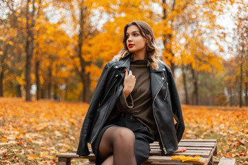 Beautiful young girl with red lips in fashionable casual clothes with a sweater, leather jacket and shorts sits and rest on a wooden pallet in an golden colorful autumn park