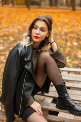 Fototapeta na wymiar Stylish beautiful young girl in fashionable casual clothes with a leather jacket, pullover and shorts sits on a wooden pallet in an autumn park