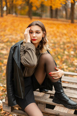 Fashionable young beautiful woman in stylish autumn casual outfit with black leather jacket, green pullover and shorts with boots sits on wooden pallet in golden fall park with yellow leaf