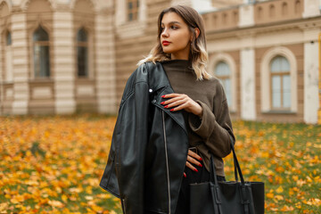 Fototapeta premium Stylish beautiful young woman model in fashionable clothes with a leather jacket and a green pullover with a leather bag walks in the autumn city with yellow foliage