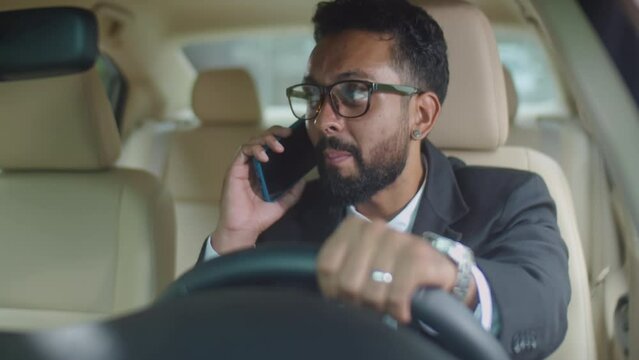 Businessman in formal outfit sitting on driver seat in car, holding steering wheel and speaking on mobile phone