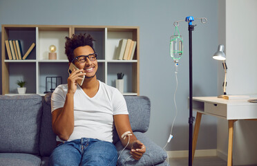 Happy, smiling young African American man talking with doctor on cell phone while sitting on couch at home and receiving medication infusion through intravenous drip