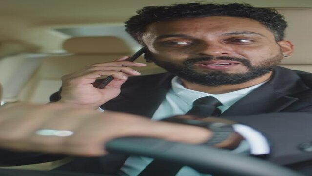 Vertical shot of professional businessman in formal suit sitting on driver seat in car, holding steering wheel and speaking on cell phone