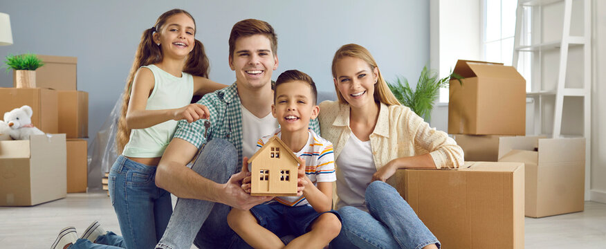 Portrait of happy young family in new home on moving day. Mom, dad and children sitting on floor in living room with cardboard boxes, holding toy house and smiling. Buying real estate property concept