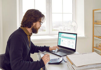 Busy man working in his business office. Young businessman sitting at office desk, using modern laptop computer, looking at online banking website page, and entering some data