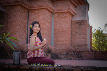 Beautiful thai woman is sitting in an old temple smiling and looking to the camera while holding a frangipani flower in her hand next to her have tiffin.