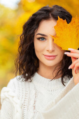 Autumn fresh portrait of a beautiful young woman with a clean face, curly hair and blue eyes in a fashion vintage white knitted sweater covers face with an autumn maple yellow leaf in the park.