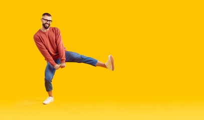 Fototapeta na wymiar Funny young man in modern outfit dancing on copy space studio background. Happy guy in orange sweatshirt, blue jeans and glasses dancing isolated on yellow copyspace background. Casual fashion concept