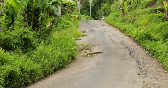 Man steer motorbike around potholes at rural road, rides towards and passes. Asphalt weathered and affected badly by water flows, crumbling fast at inclined part of street. Green tropical plants