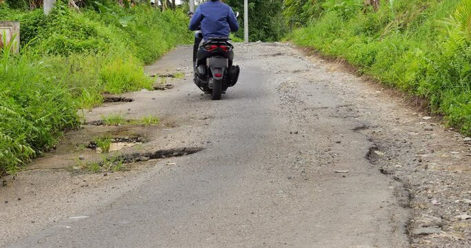 Motorbike ride around pothole but then ride over other one. Damaged road surface at Bali countryside, due to heavy rains and bad drainage at this place